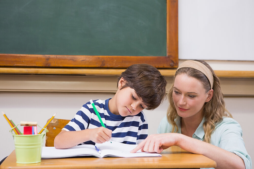 Schulbegleitung erklärt jungem Schüler eine Aufgabe, die dieser gerade bearbeitet